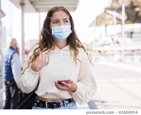 Woman in mask holding smartphone while waiting for tram Woman in mask holding smartphone while waiting for tram 81600054