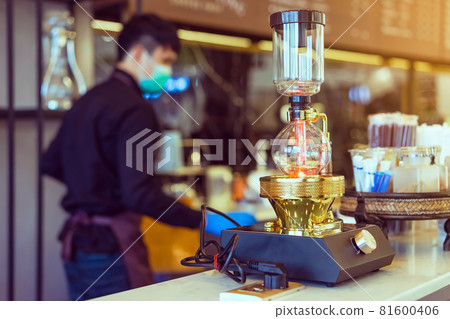 Close up to barista make coffee with Vacuum Pot Syphon Brewer. Syphon (Siphon) coffee maker brews drink using two chambers where vapor pressure and vacuum produce coffee in cafe. Selective focus. Close up to barista make coffee with Vacuum Pot Syphon Brewer. Syphon (Siphon) coffee maker brews drink using two chambers where vapor pressure and vacuum produce coffee in cafe. Selective focus. 81600406
