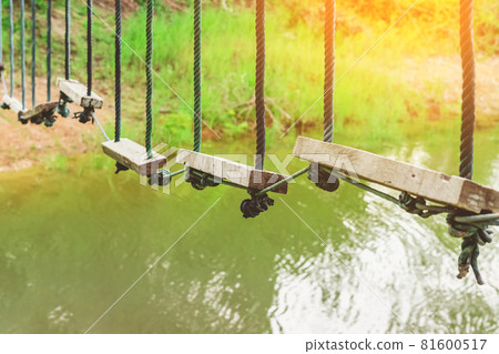 Many swings made of wooden board and tied with rope hang on cable over a small stream flowing through a tropical green rainforest. Swing hanging high and frightening above the river. Selective focus. Many swings made of wooden board and tied with rope hang on cable over a small stream flowing through a tropical green rainforest. Swing hanging high and frightening above the river. Selective focus. 81600517