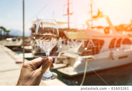 Glass of wine in hand. A glass of white wine against the backdrop of the Mediterranean sea and the port with yachts in a tourist town in the summer under sunlight. Summer, travel, lifestyle Glass of wine in hand. A glass of white wine against the backdrop of the Mediterranean sea and the port with yachts in a tourist town in the summer under sunlight. Summer, travel, lifestyle 81603261