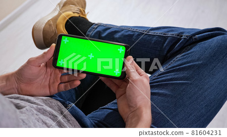 Unrecognizable man sitting on a chair and holding a phone with a green screen Unrecognizable man sitting on a chair and holding a phone with a green screen 81604231