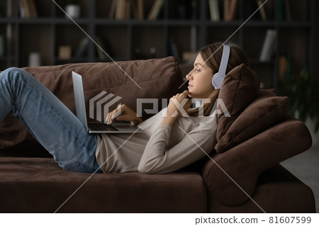 Side view woman in headphones lying on couch with laptop 81607599