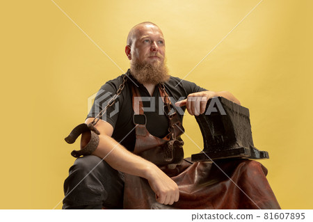 Comic portrait of muscular bearded bald man, blacksmith in leather apron or uniform isolated on yellow studio background. Concept of labor, retro professions, power, strength, humor Comic portrait of muscular bearded bald man, blacksmith in leather apron or uniform isolated on yellow studio background. Concept of labor, retro professions, power, strength, humor 81607895