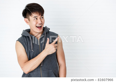 Asian handsome fit smiling man holding point the finger to copy space wearing sleeveless t-shirt gray sportswear in white background. 81607966
