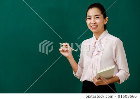 Portrait closeup shot of Asian young female beautiful school teacher tutor professor standing smiling look at camera holding pencil and textbook in hands in front of chalkboard in classroom college Portrait closeup shot of Asian young female beautiful school teacher tutor professor standing smiling look at camera holding pencil and textbook in hands in front of chalkboard in classroom college 81608660