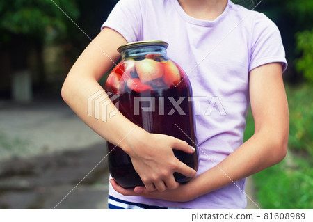 Girls holds a big jar with homemade canned apple and black currant compote on a background of green foliage 81608989