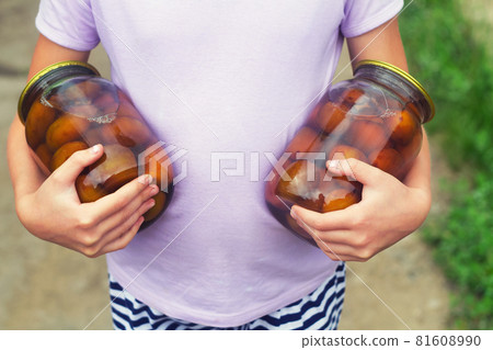 Girls holds two jars with homemade canned plum compote on a background of green foliage 81608990