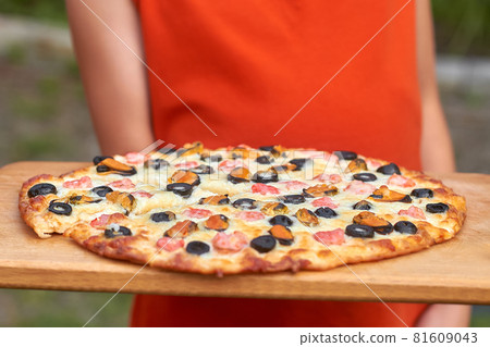 A girl in an orange t-shirt is holding homemade pizza on wooden board outdoors. Slice of pizza with shrimp, mussels, black olives and mozzarella. Home Holiday Cooking. Closeup, selective focus 81609043