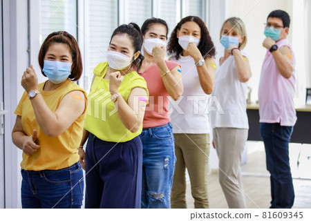 Close up shot of Asian happy strong female patient wears face mask stand look at camera in front of vaccinated orderly queue line in blurred background showing thumb up and bicep muscle up together Close up shot of Asian happy strong female patient wears face mask stand look at camera in front of vaccinated orderly queue line in blurred background showing thumb up and bicep muscle up together 81609345