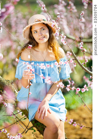Portrait of an attractive girl in blue dress and hat in blooming peach garden Portrait of an attractive girl in blue dress and hat in blooming peach garden 81609784