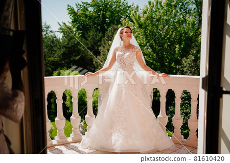 the bride in a white lace dress and a long veil stands on the terrace. 81610420