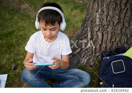 Adorable handsome elementary aged schoolboy with wireless headphones, fconcentrated on playing with smartphone, resting on the green grass of urban park, school supplies lying down next to him Adorable handsome elementary aged schoolboy with wireless headphones, fconcentrated on playing with smartphone, resting on the green grass of urban park, school supplies lying down next to him 81610492