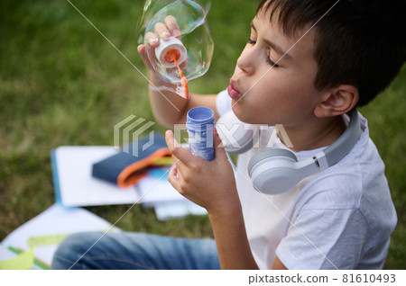 Headshot of an elementary aged schoolboy resting in the park sitting on the green grass after school, blowing bubbles. School bag with workbooks and school supplies lying down on the grass 81610493