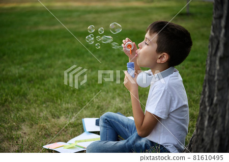 Adorable school boy enjoying his recreation between classes, blowing soap bubbles, sitting on the green grass of urban city park next to his lying down school supplies and accessories Adorable school boy enjoying his recreation between classes, blowing soap bubbles, sitting on the green grass of urban city park next to his lying down school supplies and accessories 81610495