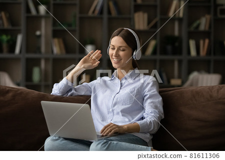 Smiling woman in headphones waving hand at webcam, using laptop Smiling woman in headphones waving hand at webcam, using laptop 81611306