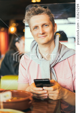 Portrait of a smiling young man using mobile phone while sitting in cafe. Make food photo for social networks, blogging, chatting, watching the video. Selective focus. Portrait of a smiling young man using mobile phone while sitting in cafe. Make food photo for social networks, blogging, chatting, watching the video. Selective focus. 81623294