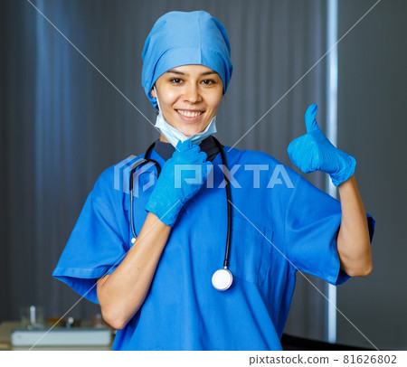 Portrait closeup shot of happy pretty female freedom doctor in blue hospital suit with stethoscope smiling thumb up when take off used surgical face mask look at camera while pandemic end 81626802