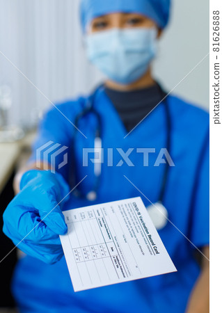 Closeup shot of Covid-19 vaccination record card certificate passport paper sent to patient by doctor wears blue hospital uniform face mask rubber gloves and stethoscope in blurred background Closeup shot of Covid-19 vaccination record card certificate passport paper sent to patient by doctor wears blue hospital uniform face mask rubber gloves and stethoscope in blurred background 81626888