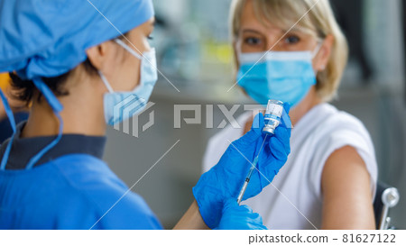 Caucasian senior patient wears face mask looking waiting to receive vaccination injection shot while doctor in blue hospital uniform preparing vaccine from glass vial dose in blurred foreground Caucasian senior patient wears face mask looking waiting to receive vaccination injection shot while doctor in blue hospital uniform preparing vaccine from glass vial dose in blurred foreground 81627122