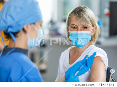 Caucasian senior patient wears face mask looking waiting to receive vaccination injection shot while doctor in blue hospital uniform preparing vaccine from glass vial dose in blurred foreground Caucasian senior patient wears face mask looking waiting to receive vaccination injection shot while doctor in blue hospital uniform preparing vaccine from glass vial dose in blurred foreground 81627124