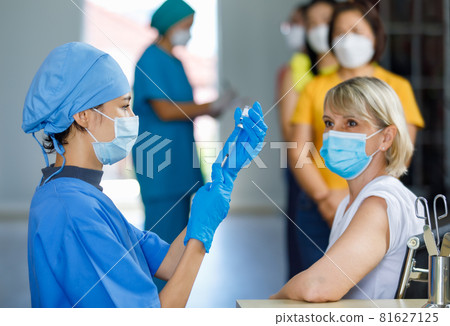 Cute doctor wears face mask rubber gloves and blue hospital uniform using needle syringe sucking coronavirus vaccine from small glass vial dose when Caucasian female patient wait for vaccination Cute doctor wears face mask rubber gloves and blue hospital uniform using needle syringe sucking coronavirus vaccine from small glass vial dose when Caucasian female patient wait for vaccination 81627125