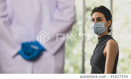 Female patient wears face mask sit wait frightened and fear while doctor in white lab coat and blue rubber gloves hold and hide vaccine syringe needle in hand behind back in blurred foreground Female patient wears face mask sit wait frightened and fear while doctor in white lab coat and blue rubber gloves hold and hide vaccine syringe needle in hand behind back in blurred foreground 81627359