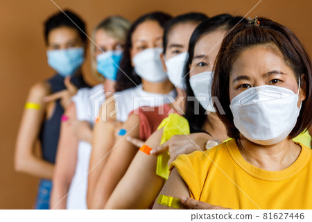 Diverse group of multinational ethnic female patients wears face mask stand in line by height order look at camera pointing colorful plasters on shoulder together after vaccinated 81627446