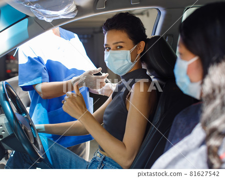 Female citizens wears face mask sitting look at camera in car with friend in drive through vaccinating line in hospital receiving coronavirus vaccine injection shot from public health doctor 81627542