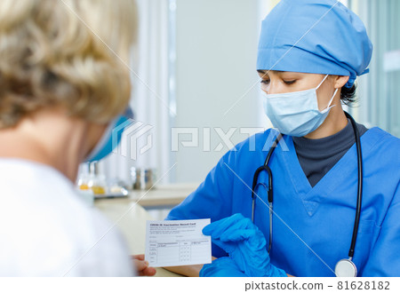Doctor in blue hospital uniform face mask and rubber gloves with stethoscope send and explain Covid-19 vaccination record card certificate to Caucasian female patient in blurred foreground Doctor in blue hospital uniform face mask and rubber gloves with stethoscope send and explain Covid-19 vaccination record card certificate to Caucasian female patient in blurred foreground 81628182