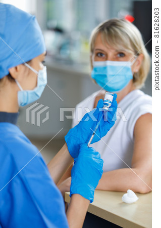 Caucasian senior patient wears face mask looking waiting to receive vaccination injection shot while doctor in blue hospital uniform preparing vaccine from glass vial dose in blurred foreground 81628203