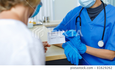 Doctor in blue hospital uniform face mask and rubber gloves with stethoscope send and explain Covid-19 vaccination record card certificate to Caucasian female patient in blurred foreground 81629262