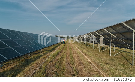 Top view of specialists walking across a solar power plant. Three employees of alternative power plant walking and talking about scheme of solar panels. Teamwork. Group of specialists. 81635379