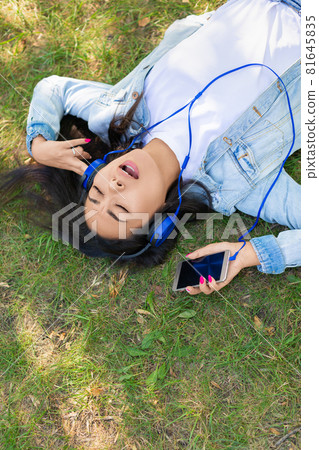 Asian woman enjoying the music while lyuing on the grass in city park - view from the top. Vertical photo. Asian woman enjoying the music while lyuing on the grass in city park - view from the top. Vertical photo. 81645835