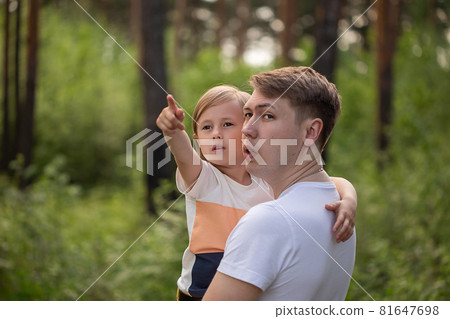 Caucasian daughter in daddy's arms points a finger to the side in the forest. Caucasian daughter in daddy's arms points a finger to the side in the forest. 81647698