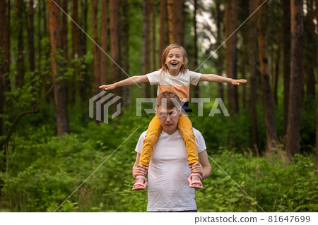 Caucasian cute little girl sitting on her father's shoulders in the forest. 81647699