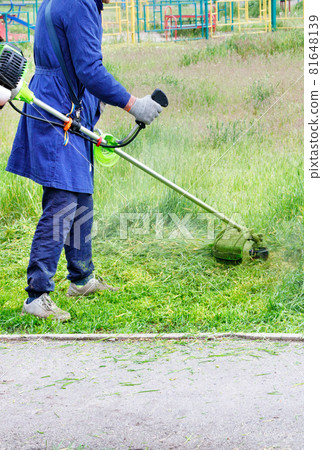 A worker in a blue overalls mows green grass with a gasoline trimmer. Vertical image. A worker in a blue overalls mows green grass with a gasoline trimmer. Vertical image. 81648139