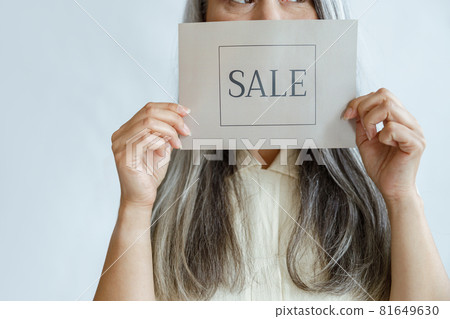 Middle aged lady with loose grey hair holds Sale sign near face standing in studio Middle aged lady with loose grey hair holds Sale sign near face standing in studio 81649630