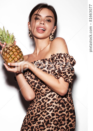 Beautiful sexy woman with pineapple in hands in a leopard dress and earrings, with classic smokey makeup and a fashionable hairstyle. Beauty face. 81663077