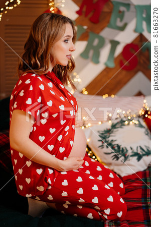 beautiful pregnant woman in red pajamas on bed in bedroom by Christmas tree. beautiful pregnant woman in red pajamas on bed in bedroom by Christmas tree. 81663270