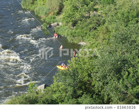 Several tourists are resting on the banks of the Ukrainian river called the Southern Bug. The bank of the Southern Bug is overgrown with trees. Fast flow of river water. 81663816