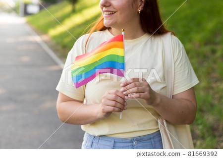 Beautiful red lesbian woman with LGBT rainbow flag at sunset on green nature background. Happiness, freedom and love concept for same sex couples. 81669392