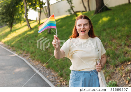 Beautiful red lesbian woman with LGBT rainbow flag at sunset on green nature background. Happiness, freedom and love concept for same sex couples. 81669397