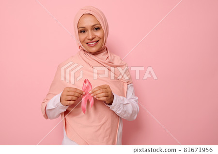 Friendly and solidar Arab Muslim woman in a pink hijab holds a pink ribbon to her chest levels for breast cancer campaign, promoting cancer awareness. Pink October month , women's health care concept 81671956