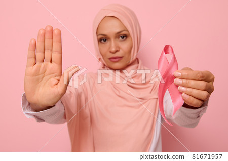 Confident portrait of blurred Arab Muslim woman in hijab holding a pink cancer awareness ribbon. Focus on woman's hand gesturing STOP , isolated over pink background with copy space. Fight Cancer Day Confident portrait of blurred Arab Muslim woman in hijab holding a pink cancer awareness ribbon. Focus on woman's hand gesturing STOP , isolated over pink background with copy space. Fight Cancer Day 81671957