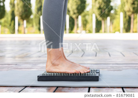 Yogi woman in gray pants standing barefoot on a wooden board with nails during outdoor yoga class Yogi woman in gray pants standing barefoot on a wooden board with nails during outdoor yoga class 81679081