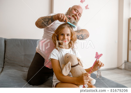Smiling girl holds toy dog and magic stick while dad brushes her hair in room Smiling girl holds toy dog and magic stick while dad brushes her hair in room 81679206