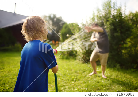 Funny little boy with his father playing with garden hose in sunny backyard. Preschooler child having fun with spray of water. Summer outdoors activity for kids. Funny little boy with his father playing with garden hose in sunny backyard. Preschooler child having fun with spray of water. Summer outdoors activity for kids. 81679995