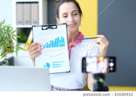 Woman blogger showing document with charts in front of mobile phone camera 81680910