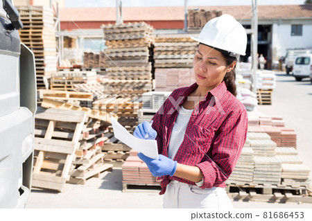 Woman in hardhat standing beside truck in outdoor warehouse 81686453
