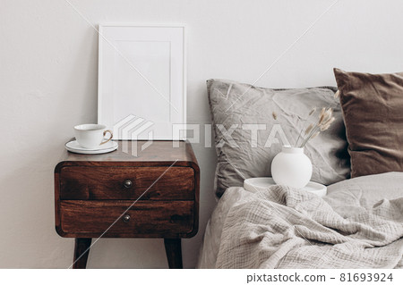 Vertical white picture frame mockup. Retro wooden bedside table. Modern white ceramic vase with dry Lagurus ovatus grass and cup of coffee. Beige linen, velvet pillows in Scandinavian bedroom. Vertical white picture frame mockup. Retro wooden bedside table. Modern white ceramic vase with dry Lagurus ovatus grass and cup of coffee. Beige linen, velvet pillows in Scandinavian bedroom. 81693924